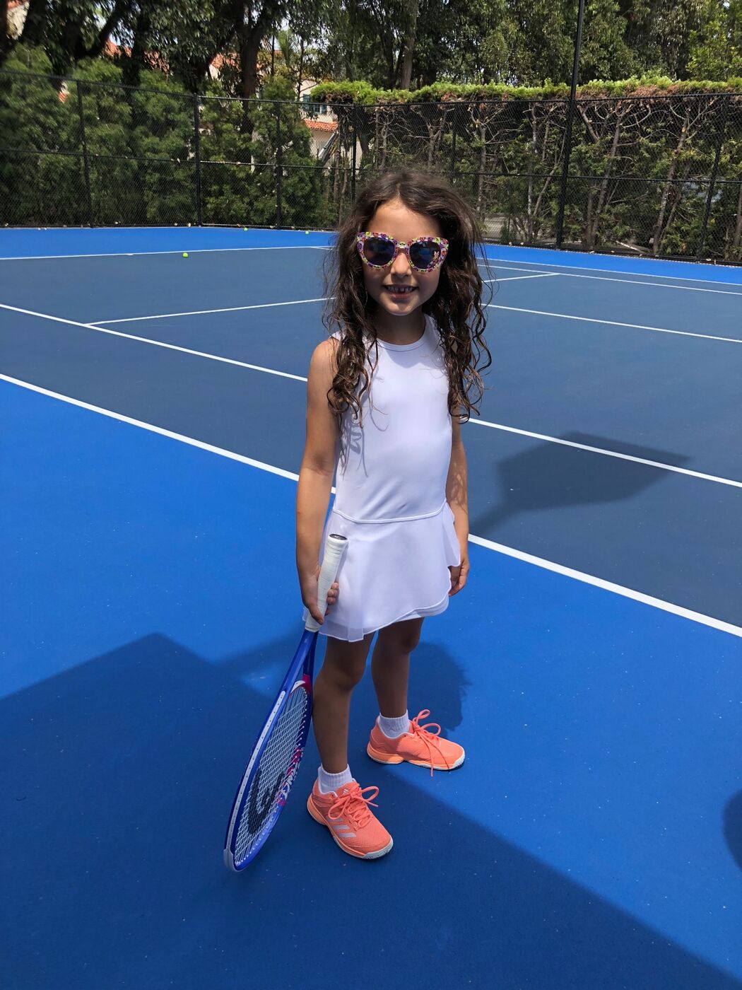 Gabi on a blue court in a white tennis dress and flower sunglasses, smiling